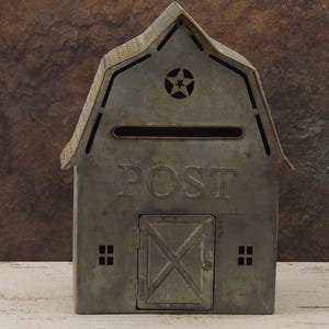 Op de afbeelding: Een metalen brievenbus in de vorm van een schuur met een stervormige uitsparing op het dak en het woord "POST" aan de voorkant. De brievenbus heeft een gleuf voor post en een kleine deur met een kruisontwerp.