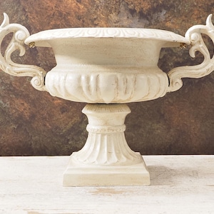 Puede incluir: Urna ornamentada de color crema con dos asas enrolladas. La urna tiene un cuenco ancho y poco profundo y se asienta sobre una base de pedestal con una parte inferior cuadrada. La superficie tiene un acabado envejecido, lo que sugiere un estilo antiguo o vintage.