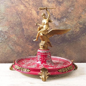 Puede incluir: Centro de mesa decorativo con una estatua dorada de una mujer montada en un águila, sosteniendo una antorcha. La estatua se asienta sobre una bandeja roja con motivos florales, detalles dorados y pies ornamentados. Altura aproximada 38 cm.