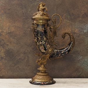 以下が含まれることがあります： 黒と花の模様のボディと金色の金属のディテールが特徴的な、装飾的なアンティークの飲用ホーン。 ホーンは蓋に馬の置物と装飾的なハンドルを備えています。 ベースは丸く、花の模様も特徴です。