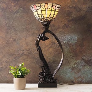 May include: A decorative table lamp with a stained glass shade in shades of yellow, pink, and white. The lamp has a dark bronze-colored base with a floral and curved design. A small potted plant sits to the left of the lamp.