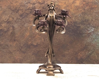 Op de afbeelding: Een bronzen kandelaar met een vrouwelijke figuur in het midden en vier kaarshouders. De figuur staat op een voet met vier poten. De kandelaar is versierd met bloemmotieven.