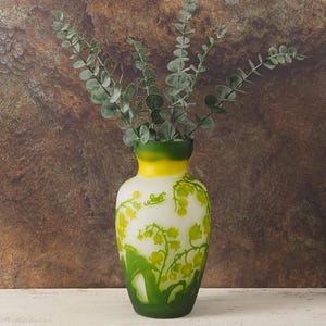 Puede incluir: Un jarr&oacute;n de cer&aacute;mica blanca con un esmalte verde y amarillo y un dise&ntilde;o floral. El jarr&oacute;n est&aacute; lleno de ramas de eucalipto verdes.
