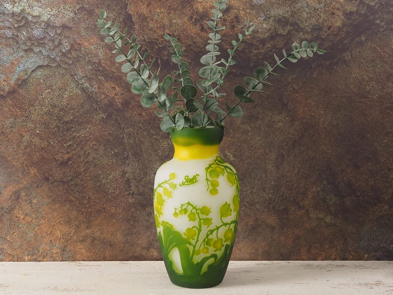 Puede incluir: Un jarr&oacute;n de cer&aacute;mica blanca con un esmalte verde y amarillo y un dise&ntilde;o floral. El jarr&oacute;n est&aacute; lleno de ramas de eucalipto verdes.