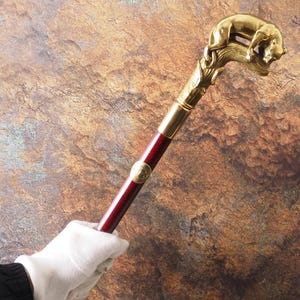 Op de afbeelding: Een decoratieve wandelstok met een goudkleurige dierenkopgreep. De stok heeft een donkerrode houten schacht met gouden accenten. De handgreep heeft een gedetailleerde sculptuur van een rustend dier. De stok wordt vastgehouden door een gehandschoende hand.