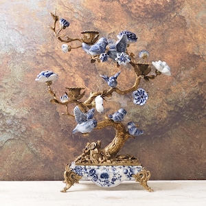 May include: An ornate candelabra in an antique style, featuring a bronze-coloured tree design with porcelain birds and flowers. The base is blue and white porcelain with a floral pattern. It has multiple candle holders and is a decorative home accent.