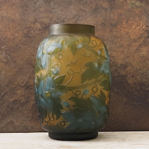 Puede incluir: Un jarrón de vidrio decorativo con un cuerpo ancho y un cuello estrecho. El jarrón presenta un diseño floral en tonos de azul y verde sobre un fondo amarillo. El jarrón tiene una base y un borde oscuros, y la palabra "Gallé" está grabada en el lateral.