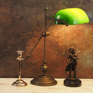 May include: An antique-style desk lamp with a green glass shade, a brass candlestick, and a bronze statue of a historical figure. The lamp has an adjustable arm and a pull chain. The statue is on a black base.