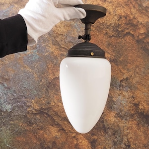 Puede incluir: Una lámpara de techo de estilo vintage con una pantalla de cristal blanca en forma de lágrima. La lámpara tiene una base de metal de color bronce oscuro y un acento de color latón. La luz es sostenida por una mano enguantada.
