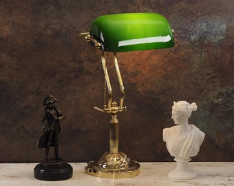 May include: A brass desk lamp with a green glass shade. The lamp has a decorative base and a chain to adjust the height of the shade.