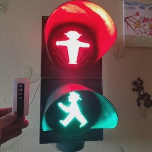 以下が含まれることがあります： 白の歩行者マークが付いた赤と緑の信号機。信号は赤で、歩行者は渡ってはいけないことを示しています。信号は、AとBの文字が書かれたリモコンで制御されています。