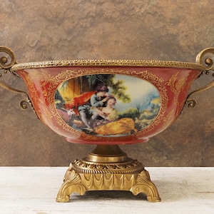 Può includere: Una ciotola decorativa antica con esterno rosso e dettagli dorati. La ciotola presenta una scena dipinta di figure in un paesaggio. Ha manici dorati ornati e poggia su una base dorata.
