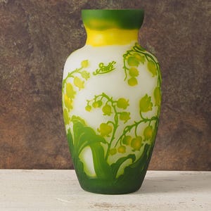 Puede incluir: Un jarr&oacute;n de vidrio blanco con un borde verde y amarillo, con un dise&ntilde;o floral de lirios del valle. El jarr&oacute;n est&aacute; firmado "Gall&eacute;".