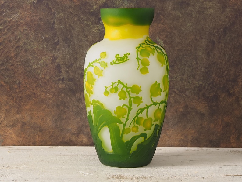 Puede incluir: Un jarr&oacute;n de vidrio blanco con un borde verde y amarillo, con un dise&ntilde;o floral de lirios del valle. El jarr&oacute;n est&aacute; firmado "Gall&eacute;".