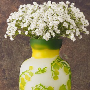 Puede incluir: Un jarr&oacute;n blanco con detalles verdes y amarillos y un dise&ntilde;o floral. El jarr&oacute;n est&aacute; lleno de un ramo de flores de aliento de beb&eacute; blancas.