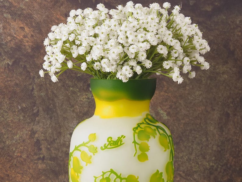 Puede incluir: Un jarr&oacute;n blanco con detalles verdes y amarillos y un dise&ntilde;o floral. El jarr&oacute;n est&aacute; lleno de un ramo de flores de aliento de beb&eacute; blancas.