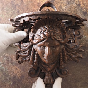 Może przedstawiać: Brązowa półka ścienna z dekoracyjną twarzą kobiety z rozwianymi włosami. Półka ma okrągły blat z ozdobnym brzegiem i zakrzywionym uchwytem u góry. Półka jest trzymana przez dłoń w rękawicy.