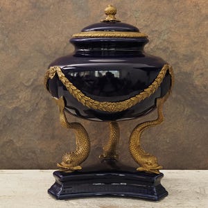 Peut inclure: Une urne à couvercle en céramique bleu marine profond ornée d'accents en métal doré, comprenant une guirlande de laurier drapée autour du corps et trois pieds sculptés en forme de dauphin reposant sur une base triangulaire bleu marine.
