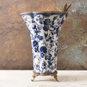 以下が含まれることがあります： 青い花柄の白い磁器製の背の高い花瓶。花瓶には金の縁と金の蝶のアクセントが付いています。3つの金の足の上に立っています。花瓶の高さは約25cmです。