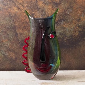 Op de afbeelding: Een decoratieve glazen vaas met een gezichtsontwerp. De vaas heeft een zwarte neus, rode lippen en een rood oog. De vaas heeft een rode, golvende accent op de zijkant en is voornamelijk groen, zwart en rood.