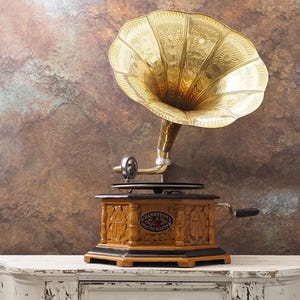 May include: A vintage gramophone with a large, gold-coloured horn. The wooden base has detailed carvings and a black trim. The gramophone is on a white, distressed surface against a textured brown background. A classic audio device.