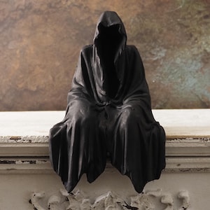May include: A black, hooded figure statue, seated with draped robes. The sculpture has a smooth, matte finish and is positioned on a distressed, white surface. The background features a mottled, brown and green wall.