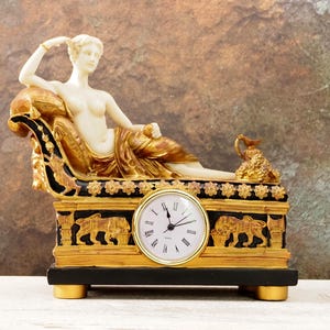 May include: A decorative clock featuring a reclining female figure in a gold and black chaise lounge. The clock has a white face with black Roman numerals and is set on a gold and black base with lion carvings.