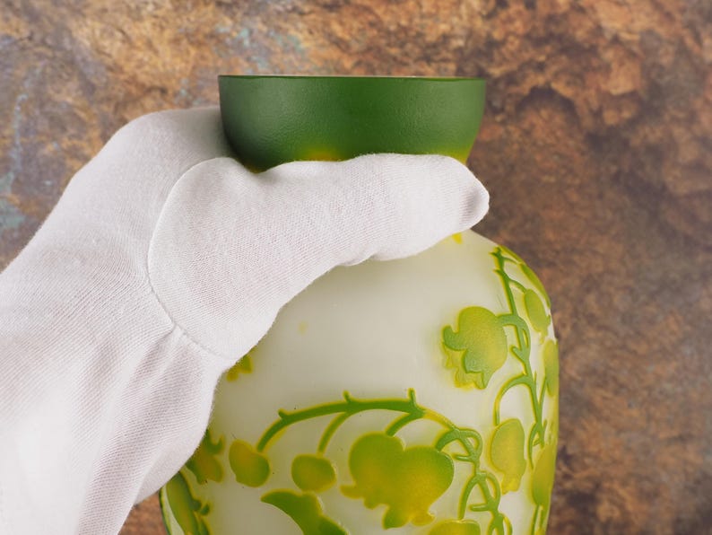 Puede incluir: Un jarr&oacute;n de vidrio blanco y verde con un dise&ntilde;o floral. El jarr&oacute;n tiene una parte superior verde y un cuerpo blanco con un dise&ntilde;o en relieve de flores verdes y amarillas.
