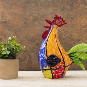 Puede incluir: Una colorida figura de gallo de vidrio con cresta roja, cuerpo naranja y azul y plumas de cola verdes. El gallo está sobre una superficie blanca junto a una pequeña planta en maceta con hojas verdes.