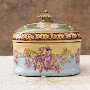 May include: An antique ceramic trinket box with a lid. The box features a light blue base with a floral design in pink and purple. The lid is green with gold accents and a gold knob. The box is a decorative item.
