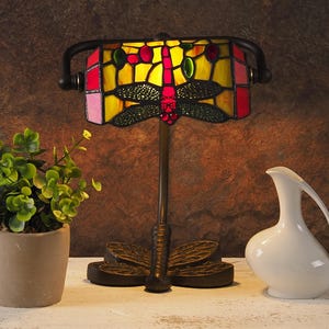 May include: A stained glass table lamp with a dragonfly design. The lampshade is yellow, red, and green with a black metal base. The base is shaped like a dragonfly.
