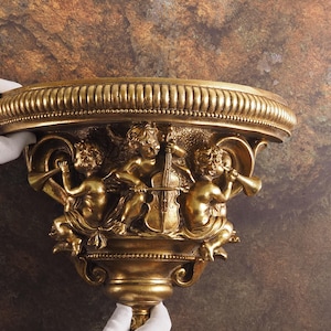 Könnte beinhalten: Ein goldfarbenes Wandregal mit einem dekorativen Design, das drei Cherubim zeigt, die Musikinstrumente spielen. Das Regal ist gebogen und hat einen gewellten Rand.