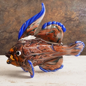Op de afbeelding: Een decoratieve glazen vissculptuur met een bruin, blauw en oranje wervelpatroon. De vis heeft grote vinnen en een gedetailleerd ontwerp, met een wit oog en een gele accent op de mond. De sculptuur staat op een wit oppervlak.