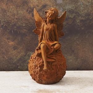 Puede incluir: Estatua de hada marrón oxidado con alas detalladas y vestido. El hada está sentada sobre una esfera floral texturizada. La estatua tiene un aspecto envejecido, ideal para la decoración de jardines. Mide unos 20 cm.