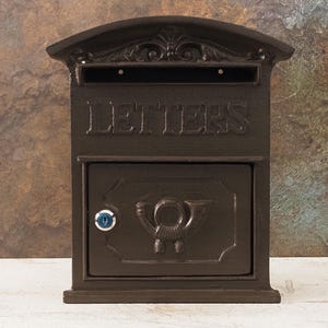 Könnte beinhalten: Dunkelbrauner Briefkasten aus Gusseisen mit dem geprägten Wort „LETTERS“ auf der Vorderseite. Der Briefkasten hat eine dekorative Oberseite und eine untere Tür mit einem Schlüsselschloss und einem Hornemblem. Für den Außenbereich konzipiert.