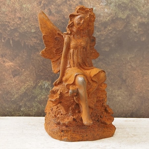 Puede incluir: Estatua de hada desgastada, con alas extendidas, sentada sobre una base texturizada. El hada está representada en posición sentada, vestida con un vestido y tiene una corona. La estatua es de un material marrón rojizo.