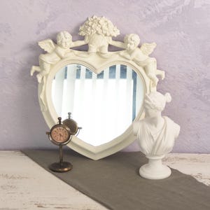 May include: White heart-shaped mirror with cherub angels on the top and sides. The mirror is surrounded by a decorative frame. A small clock and a white bust are in front of the mirror.