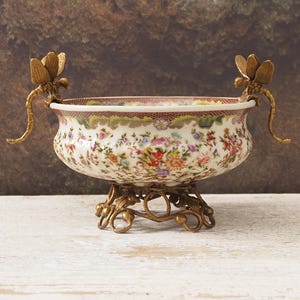 Könnte beinhalten: Eine dekorative Porzellanschale mit Blumenmuster und bronzenen Libellen-Griffen. Die Schale steht auf einem bronzenen Filigranfuß. Der Rand hat eine bordeauxrote und grüne Bordüre.