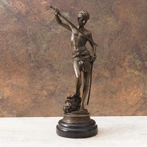 May include: Bronze statue of a young person holding a sword aloft, standing on a lion's head. The figure is draped in fabric around the waist and stands on a round base. The statue has a dark brown patina and is set against a mottled brown background.