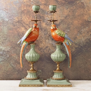 May include: Pair of decorative candle holders featuring parrot sculptures. The parrots are perched atop green and gold pedestals. The birds have orange, green, and blue plumage. The candle holders are approximately 14 inches tall.