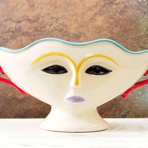 May include: A cream-colored ceramic face vase with a stylized face design. The vase features black eyes, yellow eyebrows, a lavender mouth, and red handles. The rim is outlined in light blue.