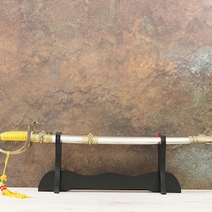 Op de afbeelding: Een decoratief zwaard met een zilveren lemmet en gouden accenten, rustend op een zwarte standaard. De greep is geel en een kwast hangt aan het handvat. Het zwaard is ongeveer 76 cm lang.