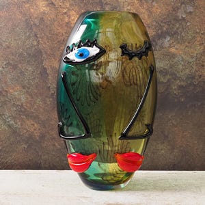 Puede incluir: Un jarrón decorativo de vidrio con diseño de rostro. El jarrón presenta un ojo azul, pestañas negras, cejas negras, labios rojos y líneas negras para la nariz. El jarrón es verde, amarillo y azul.