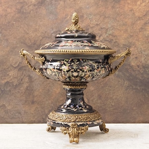 Puede incluir: Una urna decorativa negra y dorada con tapa, con motivos florales y asas ornamentadas. La urna se asienta sobre una base de pedestal con un ribete dorado detallado. La tapa está rematada con un adorno dorado.