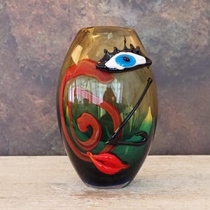 Puede incluir: Un jarrón decorativo de vidrio con diseño de rostro. El jarrón presenta un ojo azul, labios rojos y un bigote negro. El jarrón tiene una combinación de colores marrón, verde y negro.