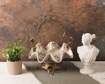 May include: A decorative bowl with a white and gold finish and a floral design. The bowl is on a stand with a gold finish and a decorative design. The bowl is in the shape of a seashell.