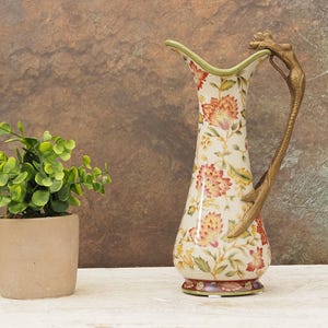 May include: A decorative ceramic pitcher with a floral pattern in red, orange, and yellow. It has a bronze-colored handle and a green rim. A small potted plant with green leaves sits to the left.