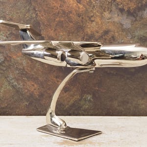 May include: A polished silver-toned metal model aeroplane featuring a sleek fuselage, two jet engines mounted under the wings, and a T-shaped tail section. The aircraft is securely attached to a curved metal stand with a rectangular base.