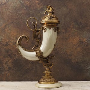 Może przedstawiać: Ozdobny, zabytkowy wazon dekoracyjny. Wazon ma kremowo-biały korpus w kształcie rogu ze skomplikowanymi, złotymi metalowymi detalami, w tym figurką konia na pokrywie i figurami przypominającymi smoki. Podstawa jest okrągła i kremowa.