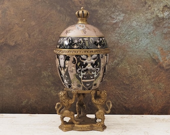May include: An ornate, egg-shaped decorative container with a crown-shaped lid finial. The container features a detailed design with black, gold, and beige accents, supported by a base with lion figures. A regal decorative piece.